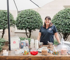 Woman behind outdoor dessert bar with candy, toppings, and cups under a white umbrella—ideal Gulf South wedding inspiration.