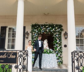 Bride and groom hold hands on stone steps outside a decorated building with floral arch, cake, and Inn sign at New Orleans wedding.
