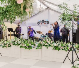 A live band plays on a flower-filled stage under a white tent, as a singer in blue performs at a Gulf South wedding.