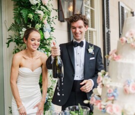 Bride and groom smile as champagne pops, with floral wedding cake and arch evoking classic New Orleans celebration.