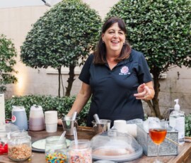 Smiling woman behind outdoor dessert table with toppings, drinks, and umbrella at a charming Louisiana wedding setting.