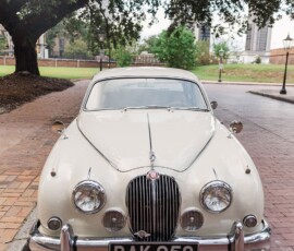 White classic car BAK 958 parked on a brick road under a tree, inspiring timeless weddings amid city buildings and lampposts.