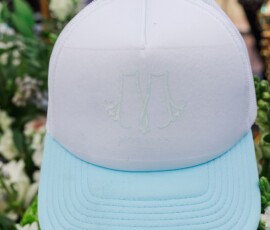 White and light blue baseball cap with subtle lettering sits among green leaves and white flowers; blurred person in background.