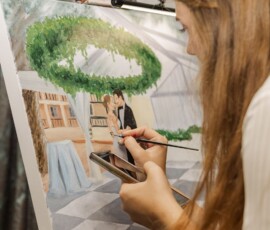 Woman with long hair paints a Gulf South wedding couple under a greenery arch, focusing on canvas details.
