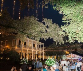 Elegant outdoor evening wedding at a large house with string lights, guests mingling among tables, flowers, trees, and a decorated tent.