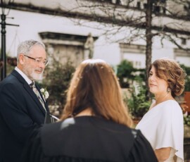A couple smiles at each other during an outdoor Gulf South wedding ceremony, officiant standing between them facing away.