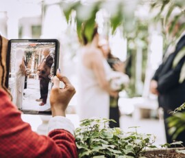 Person in red shirt records Louisiana wedding couple at altar with tablet, greenery and bright indoor setting in view.
