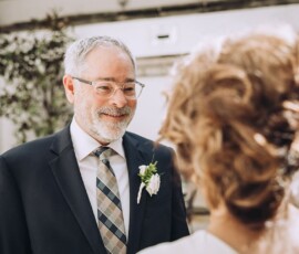 Older man in a suit with boutonniere smiles at curly-haired woman outdoors, likely at a Louisiana wedding or event.