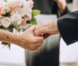 Close-up of two people holding hands at a wedding, both with rings; floral bouquet and cross tattoo add Louisiana inspiration.