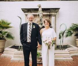 Smiling older couple in formal attire stands hand in hand by a fountain, offering charming wedding inspiration.