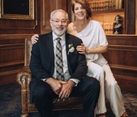 Smiling couple in formal wear pose in a wood-paneled library, surrounded by books and classic decor for wedding inspiration.