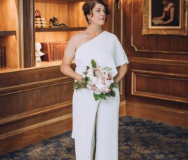 Woman in white one-shoulder jumpsuit holding flowers in wood-paneled room with bookshelves—modern wedding inspiration.