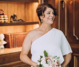 Woman in white one-shoulder dress smiling with bouquet in elegant room, Louisiana wedding atmosphere.