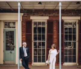 Couple outside a brick building with ornate ironwork; woman holds bouquet, man in suit, classic New Orleans wedding.