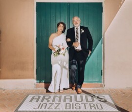 Bride in white dress and man in suit pose arm-in-arm at Arnauds Jazz Bistro entrance, sunlight highlighting Gulf South charm.
