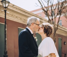 Older couple in wedding attire kisses by a pink building and leafless tree, celebrating a romantic New Orleans wedding outdoors.