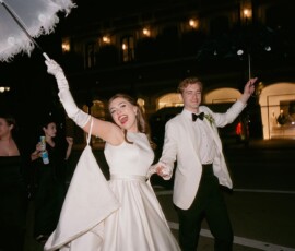 Bride and groom in formal attire parade at night, holding hands with a parasol, followed by people on a New Orleans street.