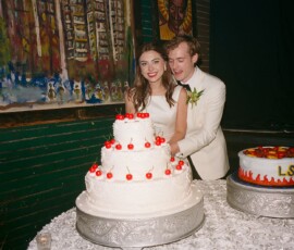 Bride and groom cut a white wedding cake with cherries at their Louisiana wedding, with LSU cake and green brick wall behind.