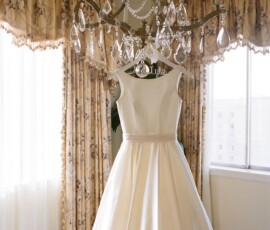 White sleeveless wedding dress hanging from a chandelier in a bright room with floral curtains and large windows.