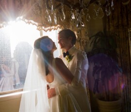 Bride and groom embrace by a sunlit window at their New Orleans wedding, with city buildings visible outside.