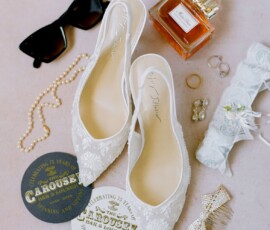 White lace bridal shoes with jewelry, perfume, and accessories arranged on a pink surface for a Gulf South wedding.
