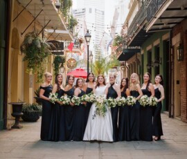 Bride in white and eleven bridesmaids in black pose with bouquets on a lantern-lit New Orleans street.
