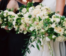 Four women in dark dresses and one in a white dress smile with large white and green bouquets at a Gulf South wedding.