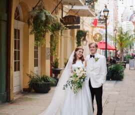 Bride in white gown and veil with groom in white tuxedo, smiling with bouquet on city street amid plants and string lights.