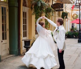 Bride and groom dancing on a city street with plants and balconies, smiling joyfully in wedding attire.