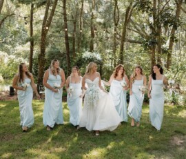 Bride in white walks outdoors with six bridesmaids in light blue, all holding bouquets amid trees at a New Orleans wedding.