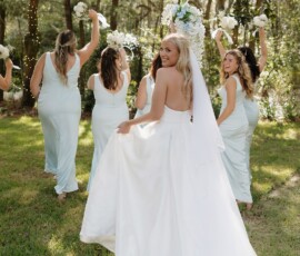 Bride in white gown smiles at the camera as bridesmaids in blue dresses stand ahead in a green, wooded New Orleans setting.