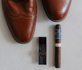 Brown dress shoes, a Happy Chic Man cologne bottle, and a dark cigar are neatly arranged on a light surface for timeless style.