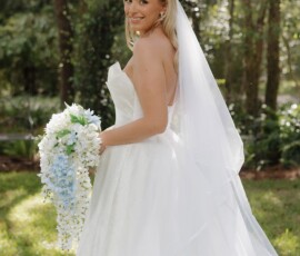 Bride in strapless white dress and veil, holding blue and white bouquet, smiles outdoors in sunlit New Orleans woods.