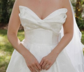 Bride in a strapless white dress with cross necklace and ring stands outdoors at her Louisiana wedding, veil and greenery behind.
