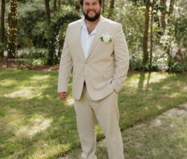 Smiling man in beige suit and white shirt stands on grass under string lights in a sunlit wooded area—Gulf South wedding inspiration.