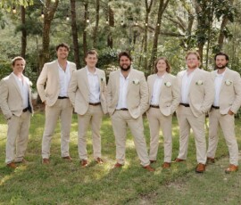 Seven men in beige suits and brown shoes pose on grass under string lights and trees, capturing Louisiana wedding inspiration.