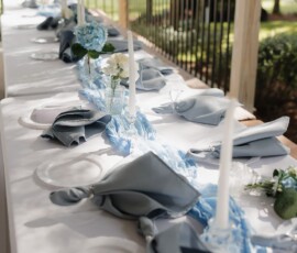 Elegant outdoor wedding table with white plates, blue napkins, candles, and flowers under sunlit trees in Louisiana.