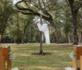 Wooden cross with white fabric between trees, pews, and flower petals on grassy aisle—romantic Gulf South wedding decor.
