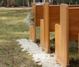 Wooden benches with white rose petals and baby’s breath, set outdoors on grass amid trees for Gulf South wedding inspiration.