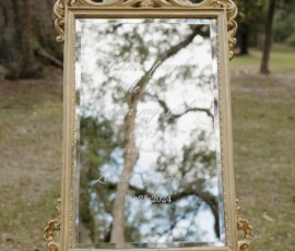 Gold-framed mirror on a wooden stand outdoors reflects trees and sky; white script displays Bible verse and Louisiana wedding details.