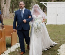 Bride in white gown and veil walks down petal-lined outdoor aisle with man in blue suit, classic Louisiana wedding backdrop.
