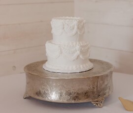 Elegant two-tier white cake with decorative icing on silver stand, displayed on a white tablecloth with wood paneling behind.