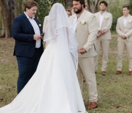 Bride and groom hold hands outdoors at their Louisiana wedding, with an officiant and beige-suited groomsmen in the background.