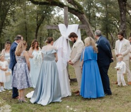 Wedding party prays outdoors in formal blue, gray, and beige by a cross, with trees surrounding this serene Louisiana setting.