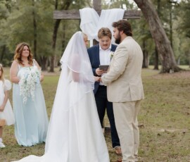 Bride and groom exchange vows by a white-draped cross outdoors, with bridesmaid and flower girl in light blue nearby, trees behind.
