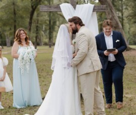Bride and groom kiss before a wooden cross at an outdoor Louisiana wedding, with attendants and lush greenery in the background.