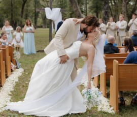 Bride and groom share a romantic dip and kiss outdoors, surrounded by guests, with trees and wooden benches in the background.