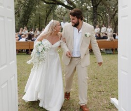 Bride and groom smiling, holding hands as they walk through white doors at an outdoor Louisiana wedding; guests seated under trees.