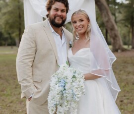 Bride in white gown and groom in beige suit smile outdoors with bouquet and wooden cross, classic Louisiana wedding.