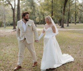 Bride in white gown and groom in tan suit hold hands, smiling as they walk through a Louisiana park wedding.